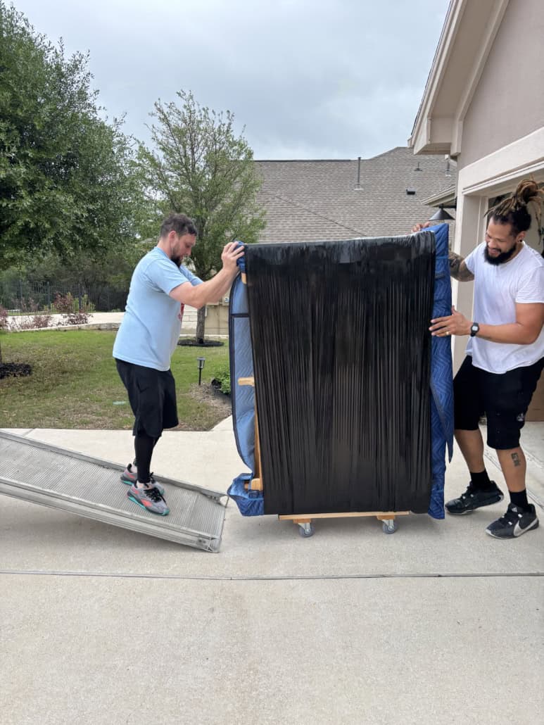 Two movers transporting a large, wrapped piece of furniture outdoors.
